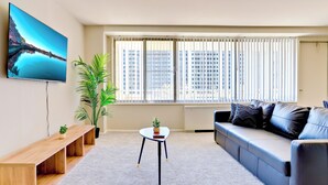 A modern living room with a large flat-screen TV, a sofa, a coffee table, and a potted plant. - Bright Comfort in DC Apts (Arlington)