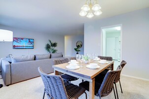 Een moderne woonkamer met een eettafel gedekt voor vier personen, een grijze bank, rieten stoelen en een flatscreen televisie aan de muur.