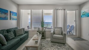Una sala de estar con un sofá verde, un sillón gris, una mesa de centro y vistas al océano a través de ventanas amplias.
