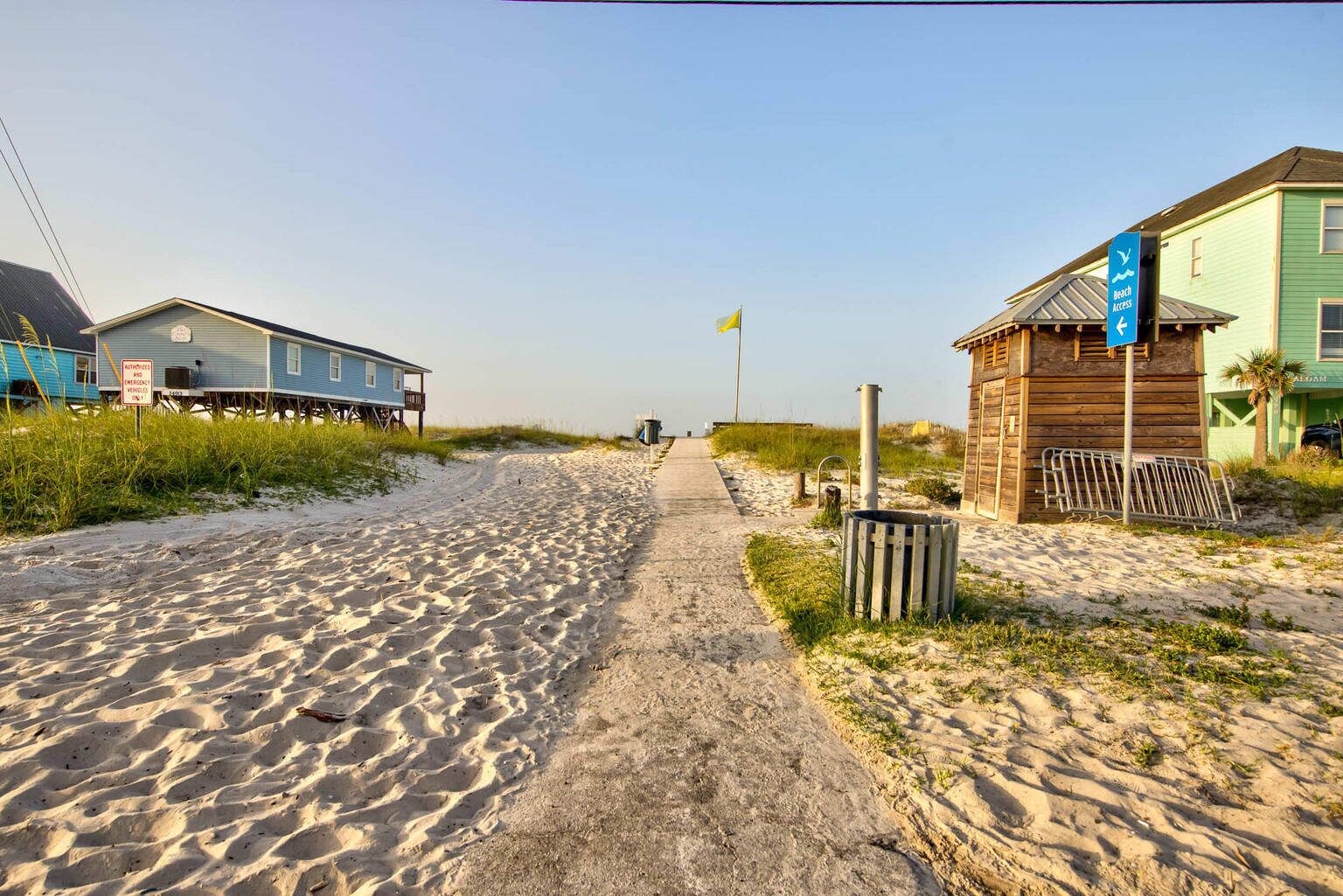 A sandy beach path leading to beach access, flanked by wooden buildings and a small wooden structure with a flag.