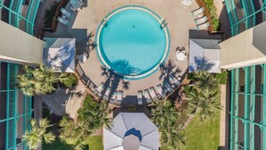 Vista aérea de un área de piscina de hotel con una piscina central, rodeada de sillas de descanso y sombrillas, y palmeras.