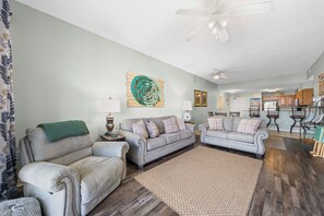 A living room with two sofas, a coffee table, a rug, a ceiling fan, and a kitchen in the background.