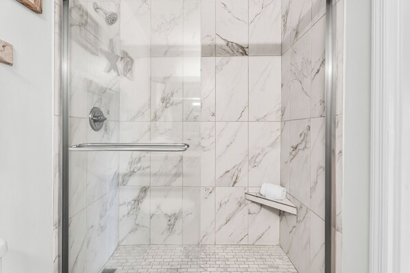 Ducha moderna con puerta de vidrio, azulejos de mármol y un pequeño estante con una toalla.