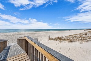 Una pasarela de madera que lleva a una playa de arena, con un cielo azul despejado y el océano al fondo.