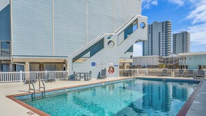Una piscina con borde de azulejos azules, un puesto de socorrista y una zona de terraza con sillas y mesas.