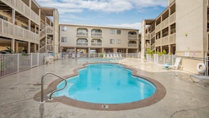 A swimming pool surrounded by a multi-story building with balconies, outdoor seating, and a clear sky.