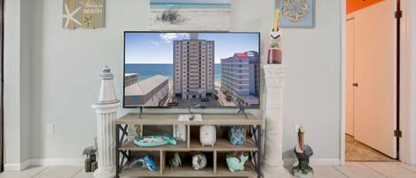 A living room with a flat-screen TV displaying a beach scene, a small shelf with decorative items, and a door leading to another room.