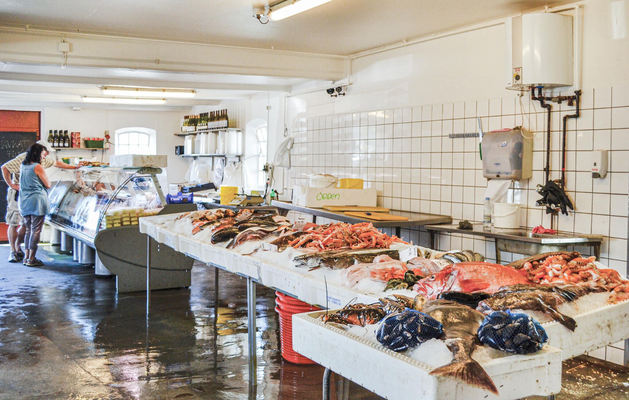 Esposizione di pesce e frutti di mare con diverse varietà, un bancone con una cassa e una persona che serve i clienti.