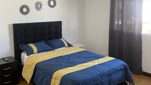 Una cama bien hecha con colcha azul y amarilla, cabecera negra y mesita de noche con control remoto.
