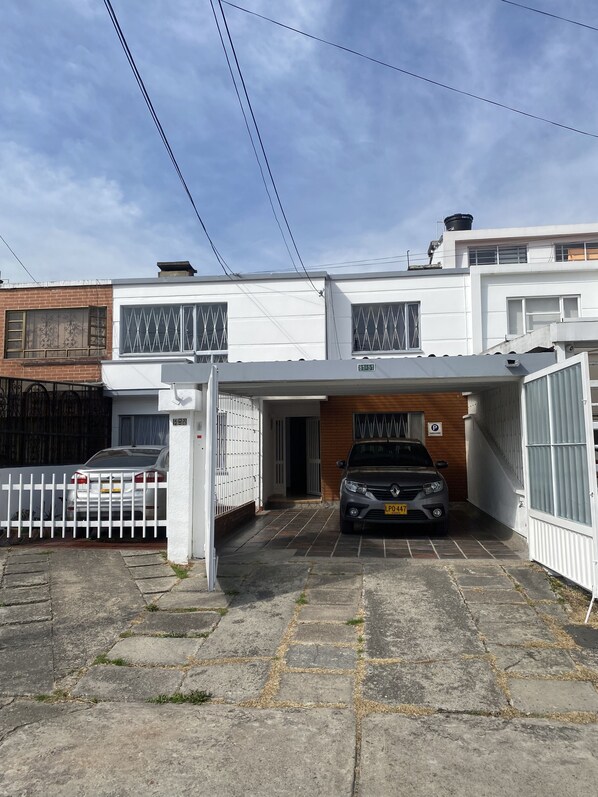 A two-story house with a white exterior, a garage with a parked car, and a gated driveway. - Hostel Urban City Bogota (Bogotá)