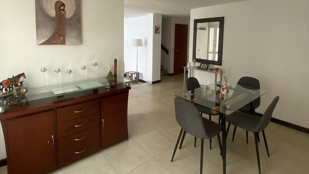 Un área de comedor moderna con una mesa de vidrio y sillas, un mueble de madera con cajones y un cuadro en la pared.