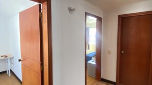 Un pasillo con suelo de madera, paredes blancas y una puerta que da a un dormitorio con cama y ventana.