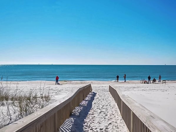 A beach with a wooden boardwalk leading to the water, clear skies, and a few people in the distance.