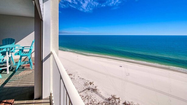 Un balcon avec des chaises turquoise donnant sur une plage aux eaux d’un bleu limpide et au rivage sablonneux.