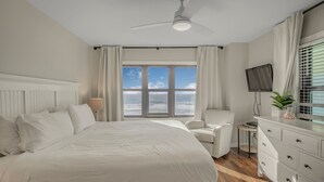 A hotel room with a large bed, a ceiling fan, a TV, a small table, a chair, and a view of the ocean. - 🌞Beachfront Condo w/Two Pools-Harbour Place-413🌞 (Orange Beach)