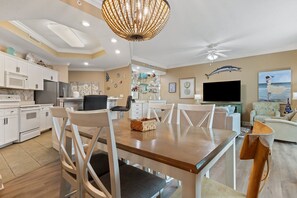 Une cuisine moderne avec des meubles blancs, un coin repas doté d’une table et de chaises en bois, un ventilateur de plafond et un salon avec une télévision et des œuvres d’art décoratives au mur.