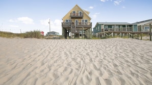 A beachfront house with a balcony and a deck, surrounded by sand and vegetation.