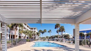 Un área junto a la piscina con sillones de descanso, una piscina, palmeras y un edificio con balcones.