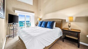 A bedroom with a large bed, a television, a nightstand with a lamp, and a view of the ocean through a window.