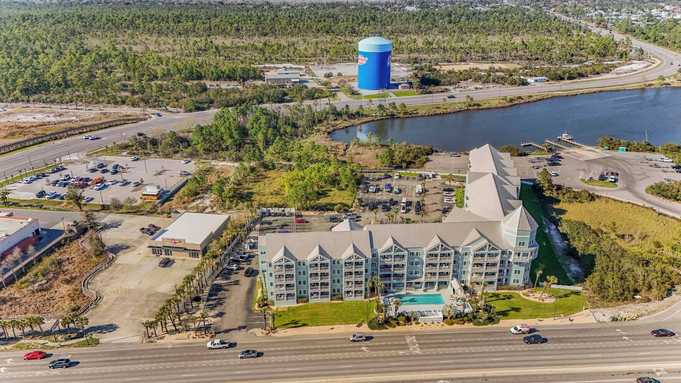 A large hotel with a central swimming pool, surrounded by a parking lot and a body of water.