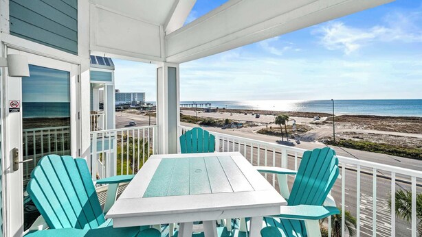 A balcony with a white table and turquoise chairs, overlooking a beach and ocean.