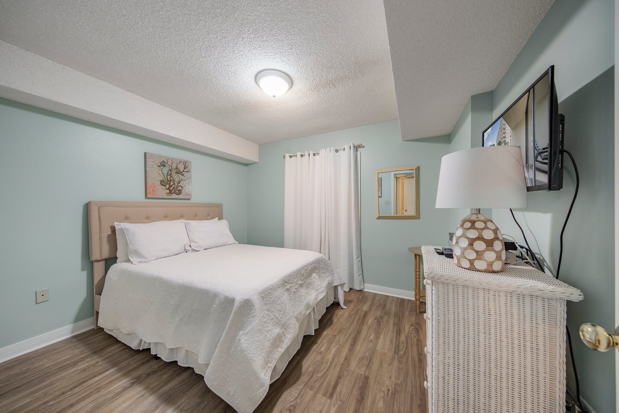 A bedroom with a bed, a bedside table with a lamp, a wall-mounted TV, and a painting on the wall.