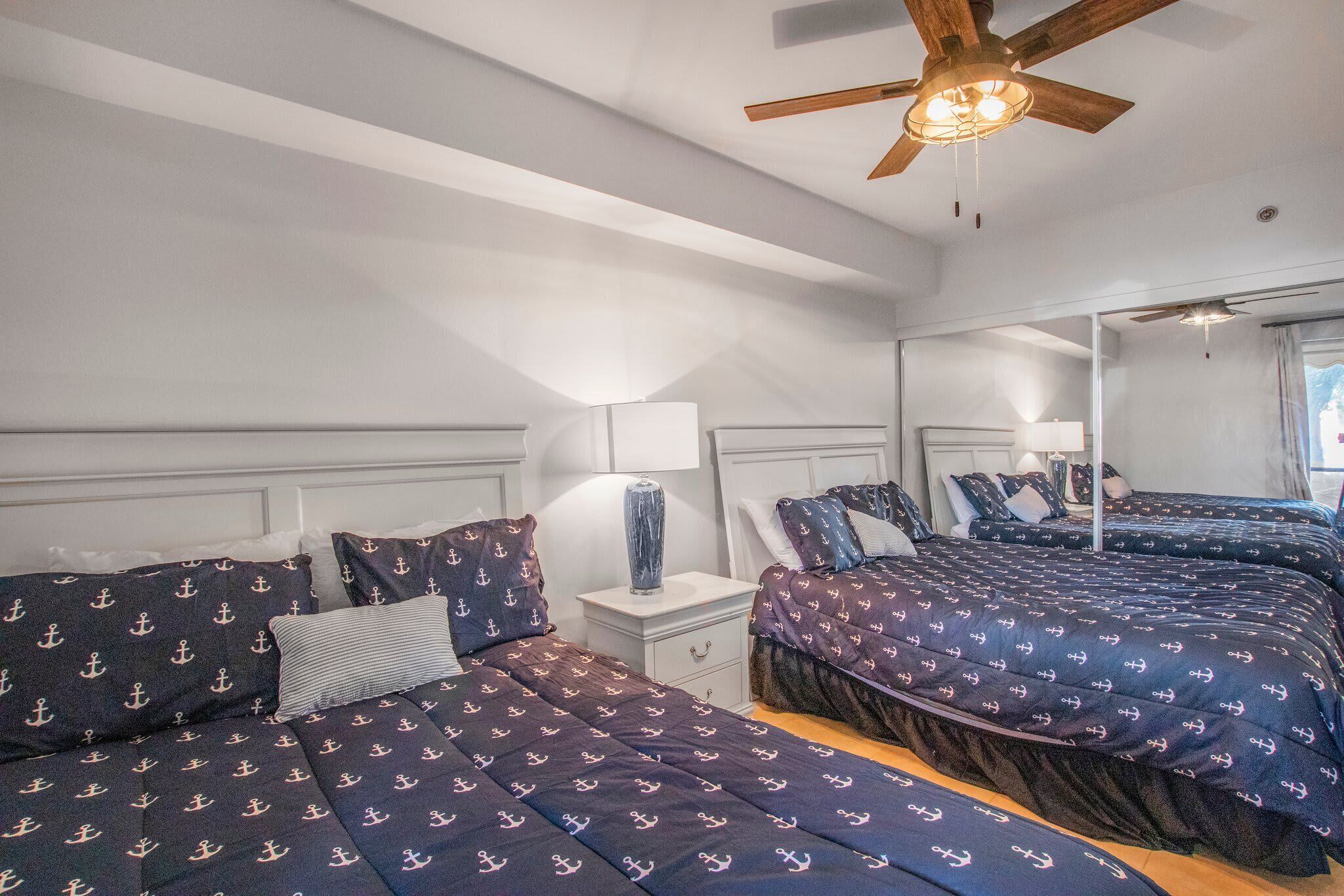 Habitación de hotel con dos camas, cada una con ropa de cama azul marino que presenta diseños de anclas. Hay un ventilador de techo, una mesita de noche con una lámpara y un espejo en la pared.