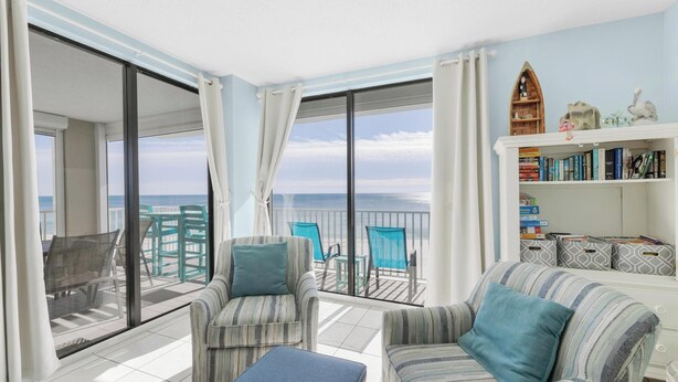 A balcony with a view of the sea, furnished with striped chairs and a sofa, and a bookshelf with books and decorative items.