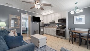 A modern kitchen with white cabinets, a dining area with a wooden table and chairs, a ceiling fan, and a TV mounted above the kitchen counter. - 🐶Pet Friendly Condo with Pool-Steps to the Beach-Sea & Sun 204🌴 (Gulf Shores)