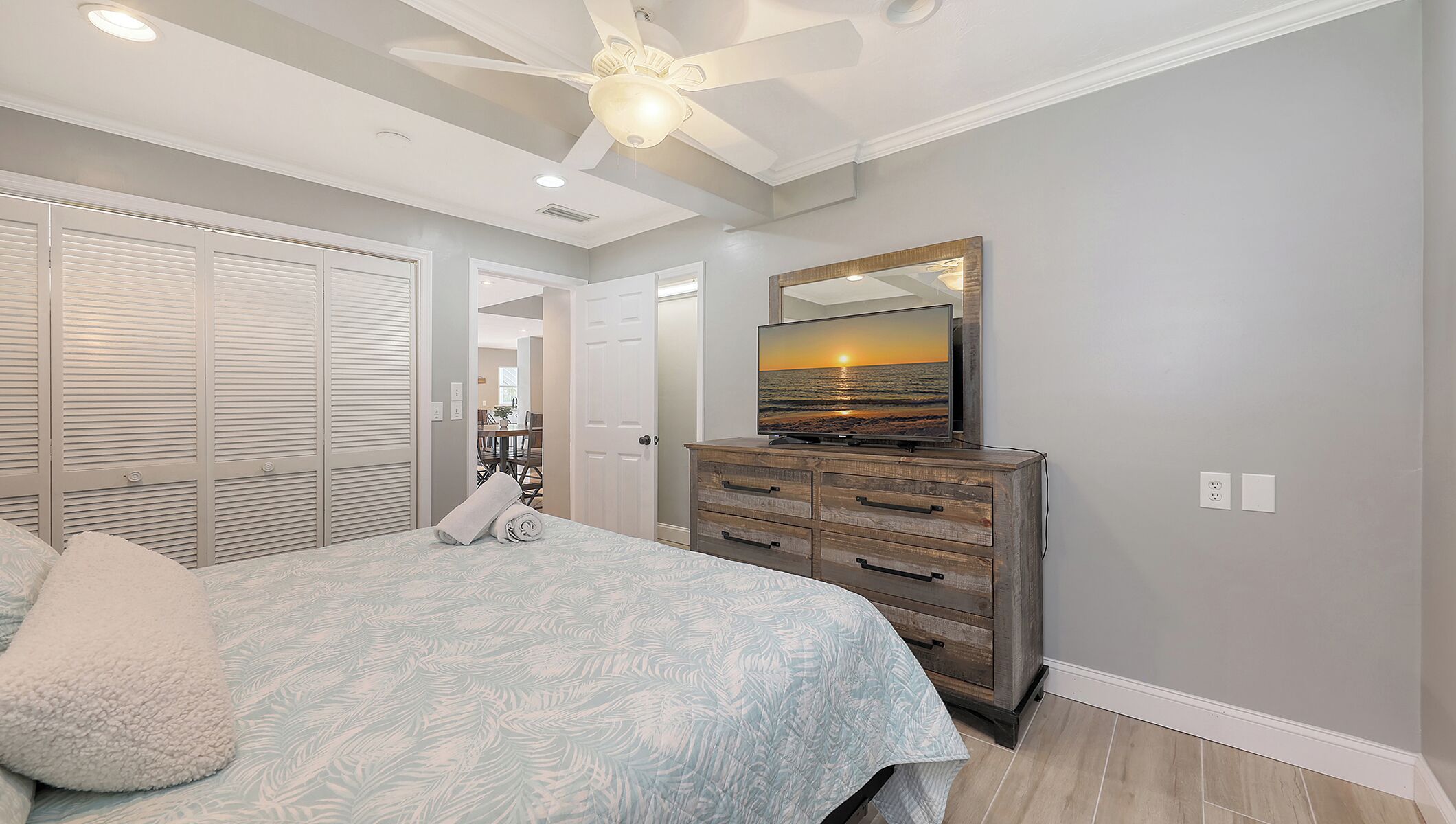 A bedroom with a bed, a dresser, a ceiling fan, and a TV displaying a sunset.