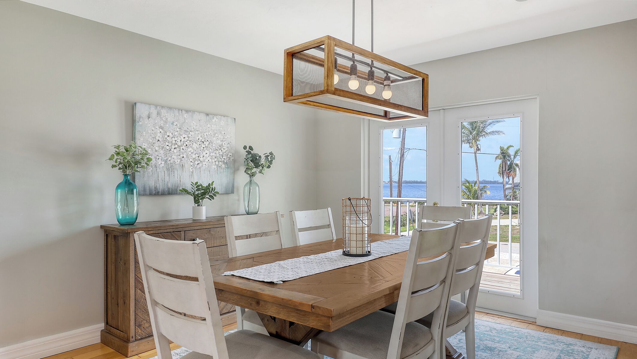 A dining area with a wooden table, white chairs, a hanging light fixture, and a view of the ocean through large windows.
