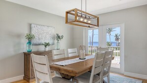 A dining area with a wooden table, white chairs, a hanging light fixture, and a view of the ocean through large windows.