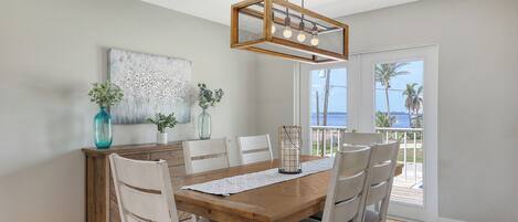 A dining area with a wooden table, white chairs, a hanging light fixture, and a view of the ocean through large windows.