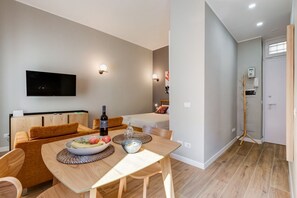 A modern living room with a flat-screen TV, wooden furniture, a dining table with fruit, and a small kitchen area.