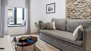 Un salón con un sofá gris, una mesita de madera con un control remoto, un cuadro en la pared y una ventana con cortinas.