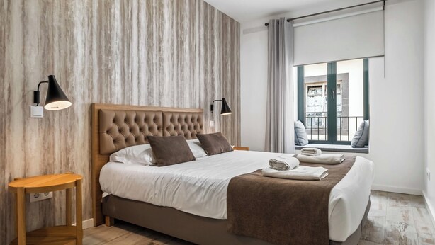 Una habitación de hotel moderna con una cama grande, cabecero de madera y vistas a la ciudad a través de amplios ventanales.