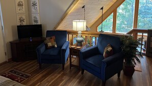 Ett mysigt sovrum på loftvåningen med ett sluttande tak, synliga träbjälkar, ett stort fönster med utsikt över träd, två blå fåtöljer, en tv och en lampa på ett träbord.
