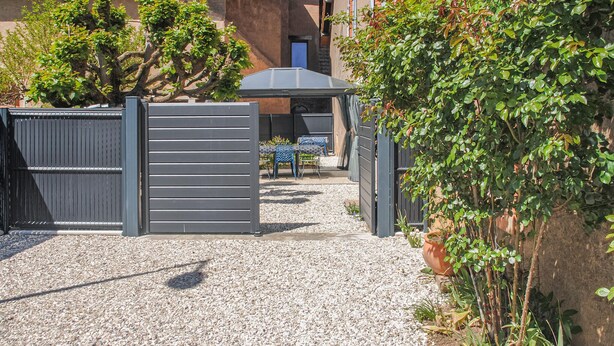 A gravel driveway leading to a gated entrance with a covered seating area and a building with a tiled roof in the background.