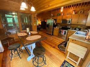 A wooden cabin interior with a kitchen area featuring a refrigerator, microwave, and stove. There are wooden tables with stools and a window with blinds.