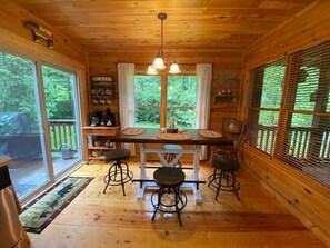 A wooden cabin interior with a dining table, bar stools, a coffee station, and large windows with blinds.
