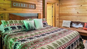 Interior de una cabaña de madera con una cama que muestra una manta con motivos de oso, un letrero que dice 'CHERRY LOC' y una zona de estar acogedora.