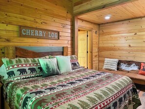 A wooden cabin interior with a bed featuring a bear-themed blanket, a sign reading 'CHERRY LOC', and a cozy seating area.