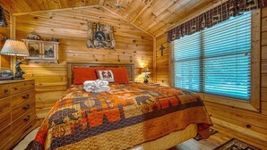 Une chambre confortable dans un chalet en rondins, avec une courtepointe matelassée, des murs en bois, une commode en bois et une fenêtre avec des stores.