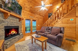 A cozy cabin interior with a stone fireplace, leather sofa, and a large window overlooking a scenic view.