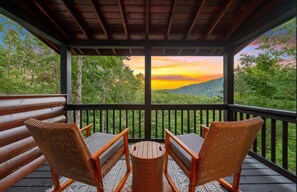 A covered outdoor seating area with two wicker chairs and a small table, overlooking a forested landscape with a sunset view.