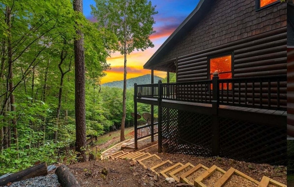 A cabin with a deck overlooking a forested area, a view of the sunset, and a satellite dish on the roof.