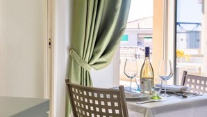 A dining table set with wine glasses and a bottle, a window with a curtain, and a chair with a lattice backrest. - Studio apartment of approximately 20 m2 composed of a living room with kitchenette equipped with 2 induction hobs, small fridge, sink,satellite TV and sofa.Sleeping area separated by sliding door with double bed, bathroom with shower without window.Air c (Marina di Cecina)