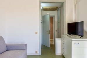 Una habitación con un sofá gris, un aparador blanco con un televisor y una puerta que da a otra habitación.