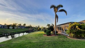 Área residencial com gramado, uma palmeira, casa com fachada de vidro e um pequeno canal.