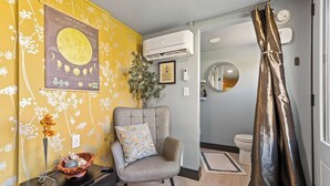 Et lille værelse med en sofa, et lille bord med en vase, en vægmonteret aircondition samt et badeværelse med toilet og bruseforhæng.